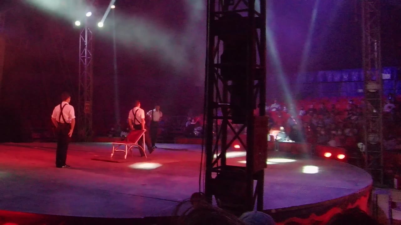 At 0:00 in San Jose Del Cabo, two male performers stand on a red-lit stage near a chair, with audience seating and stage truss visible in the background.