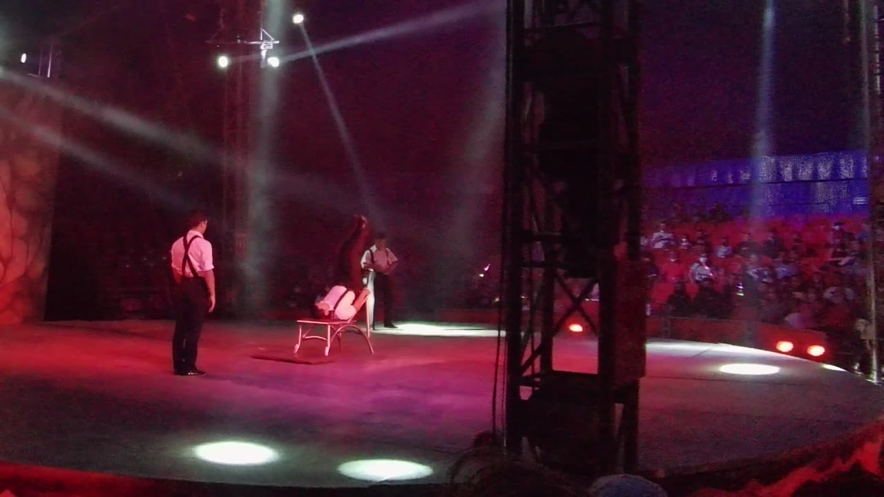Performer stands near a chair while another performer moves behind it under spotlights.