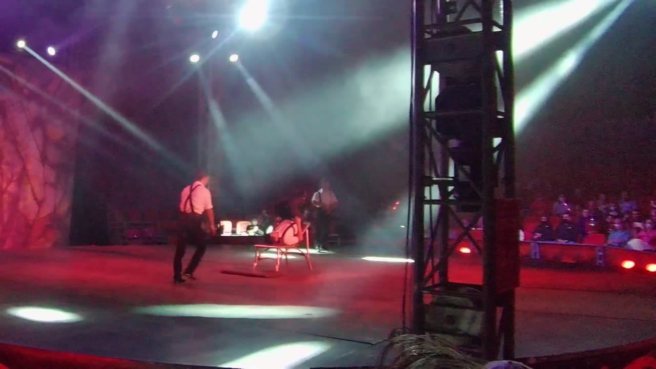 At 0:02 in San Jose Del Cabo, a performer steps toward the chair at center stage as another waits to the left, with audience rows visible beyond the lit stage.