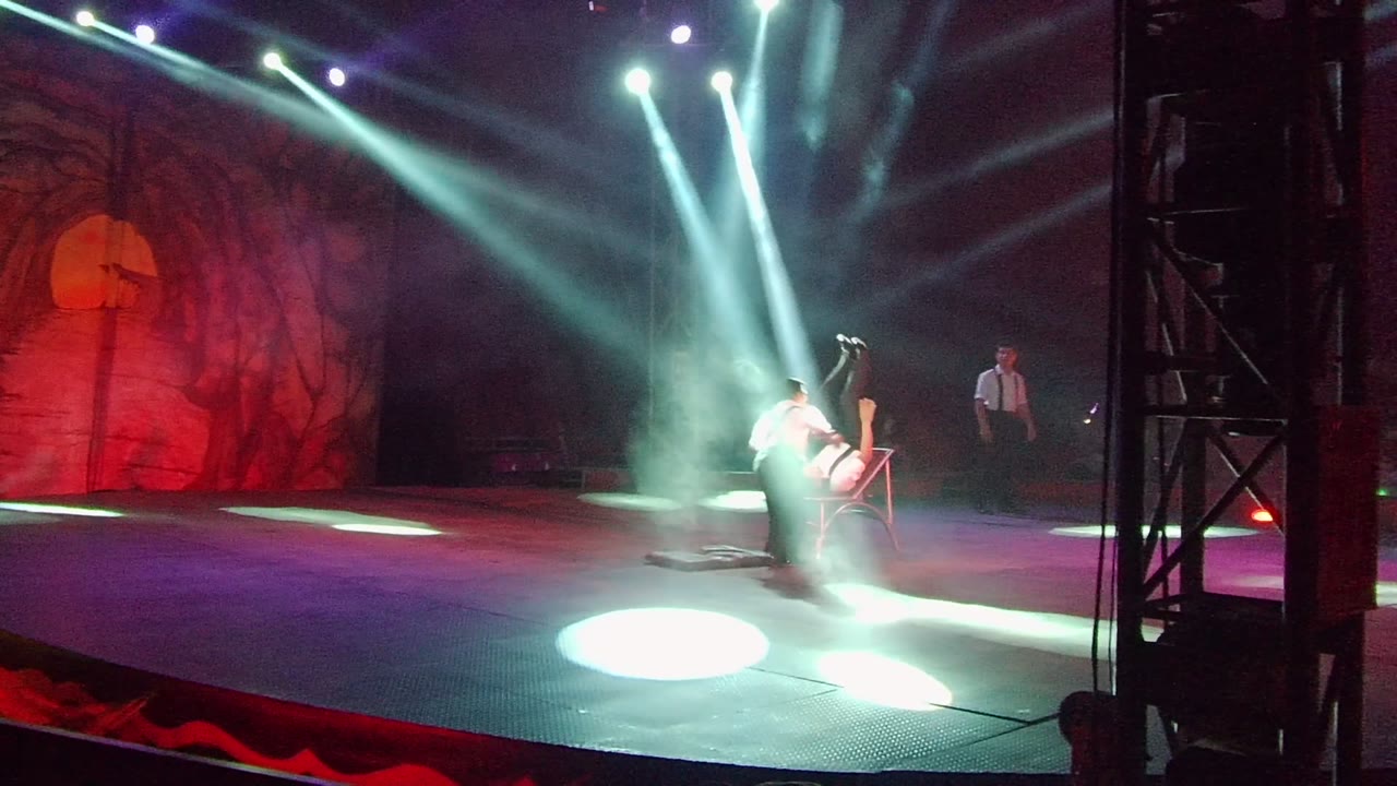Two performers in white shirts and black pants are on a lit stage in San Jose Del Cabo, Mexico, with a chair at center-right and white spotlights cutting through smoke.