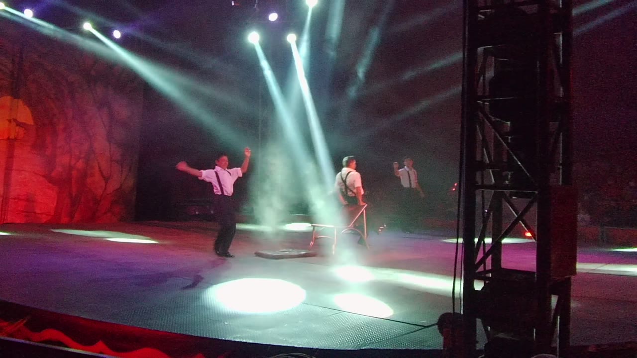 Performer gestures with arms raised while another stands by a chair.