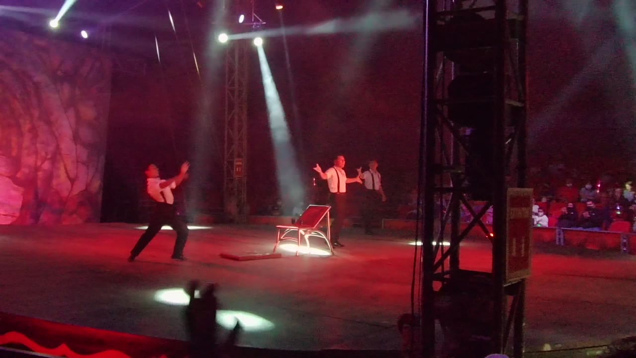 A wide view of the San Jose Del Cabo stage shows one performer moving left of the chair, another performer farther back, red wash lighting, white spotlights, and audience seating at the right edge.