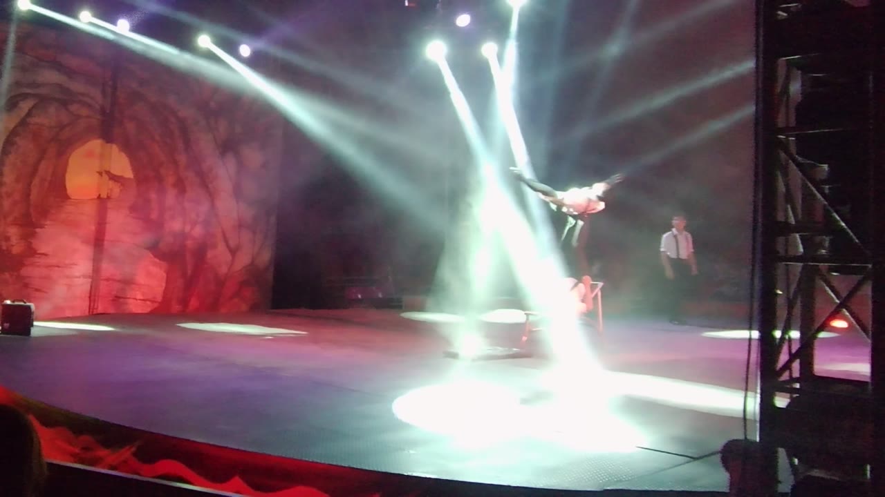 In San Jose del Cabo, an aerial acrobat stretches horizontally above a hazy stage while white spotlights cross the red cave-like backdrop and a performer in a white shirt stands at right.