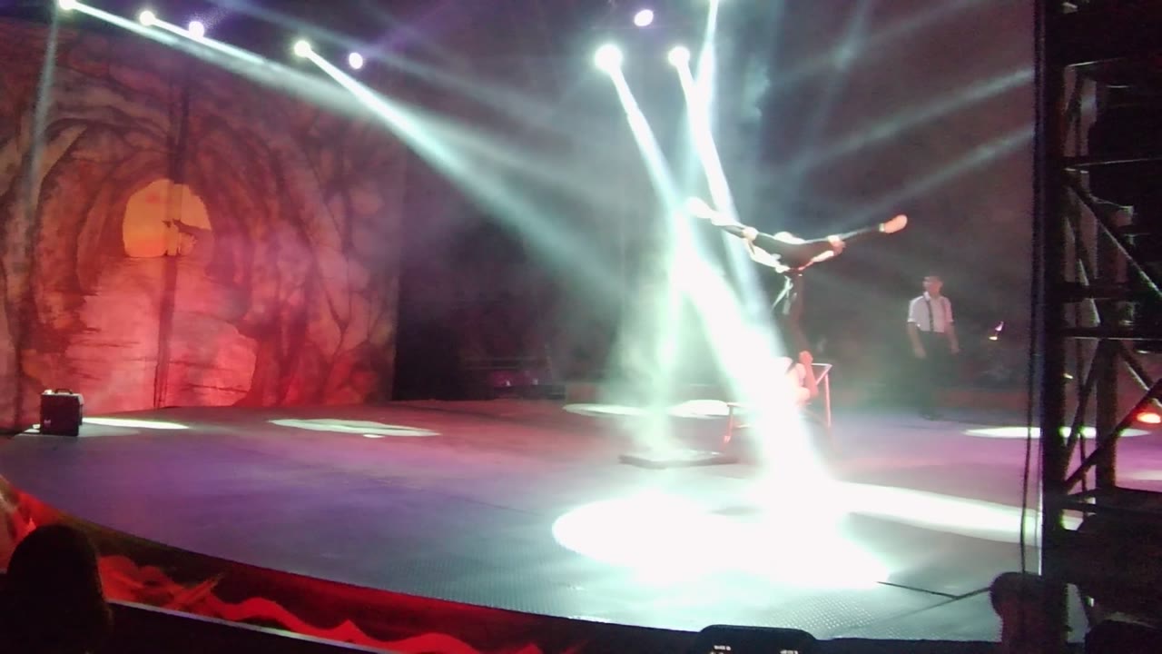 In San Jose del Cabo, the aerial performer remains high over the performance floor as intersecting white beams flare near the center of the stage and the red scenic wall fills the background.