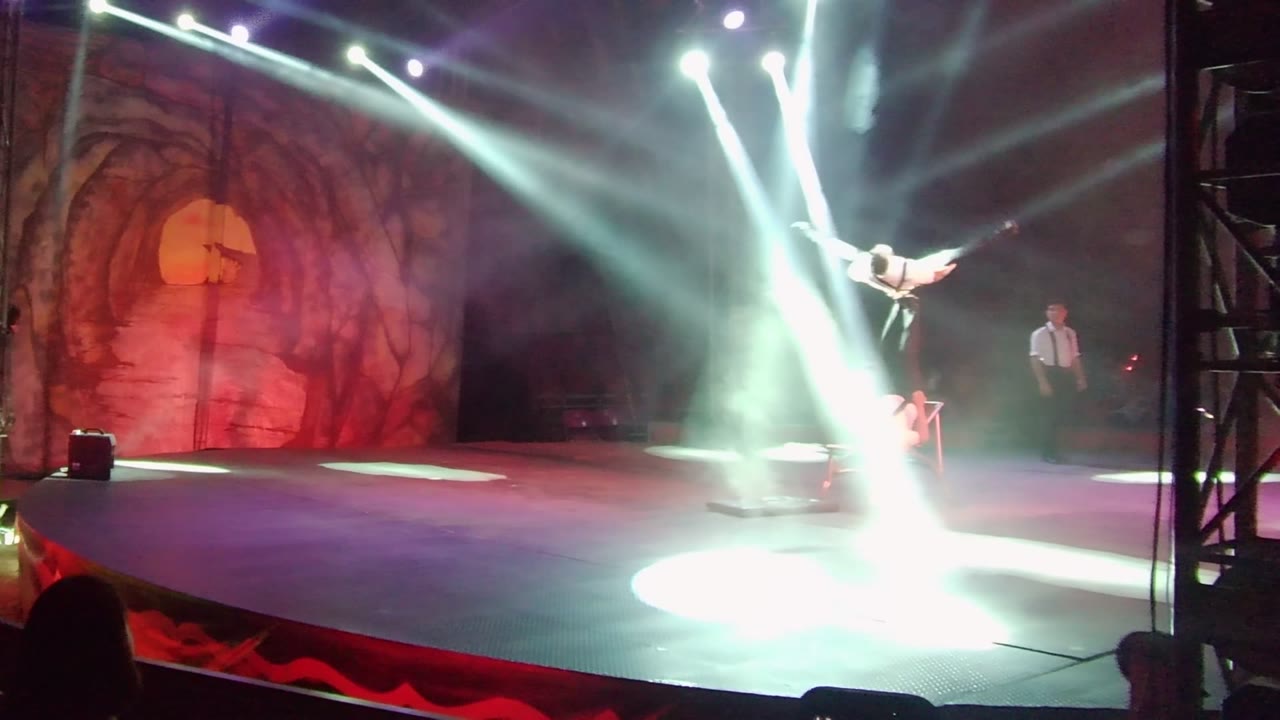In San Jose del Cabo, the aerial acrobat angles across the upper right of the stage while bright white beams wash the smoky floor and a red backdrop remains visible behind the act.