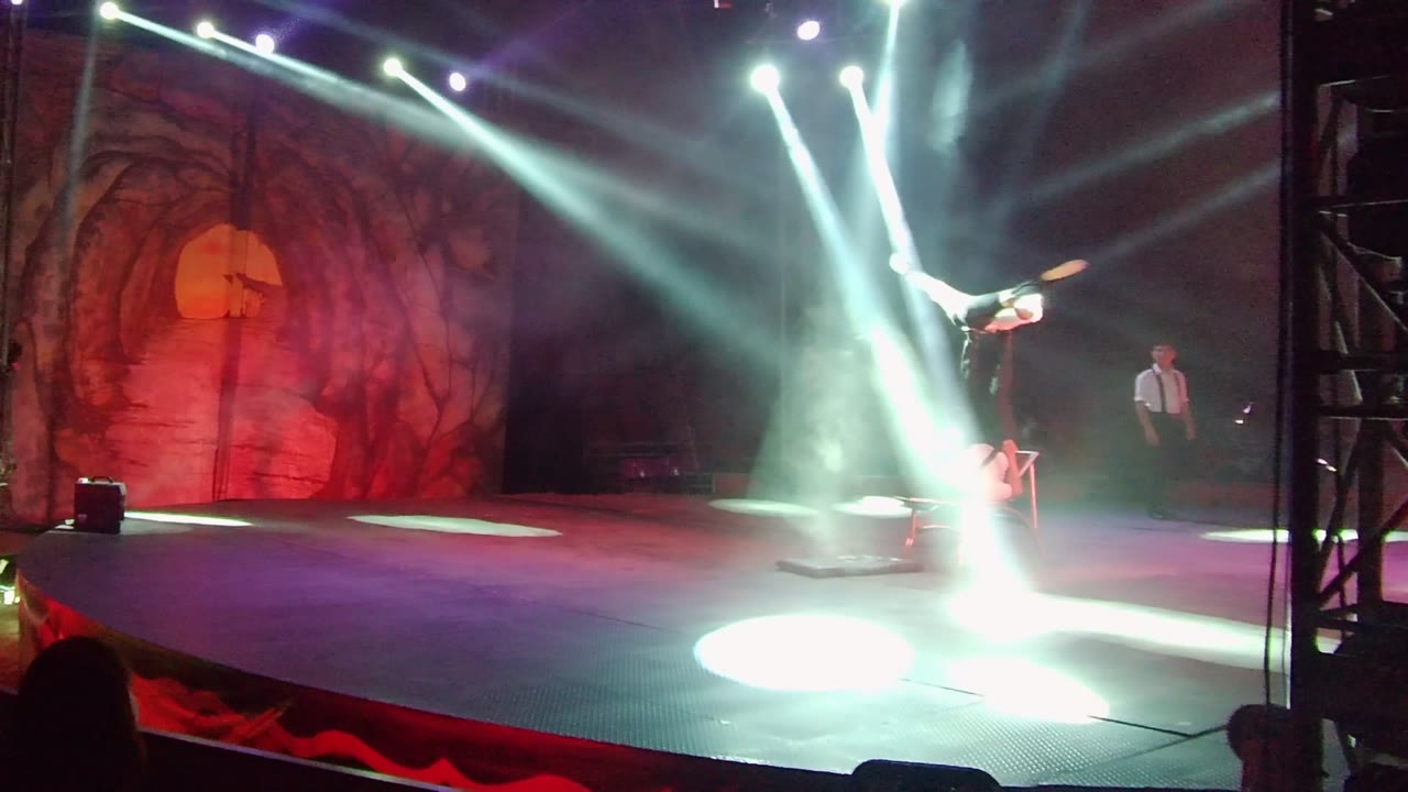 Aerial acrobat balanced near center stage under crossing spotlights.