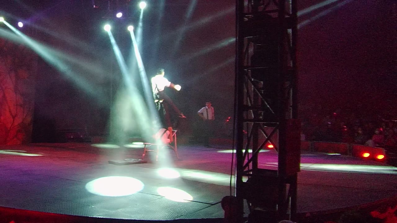 In San Jose Del Cabo, an acrobat in a white shirt is lifted above the stage while white spotlights cut through haze and audience silhouettes sit behind.
