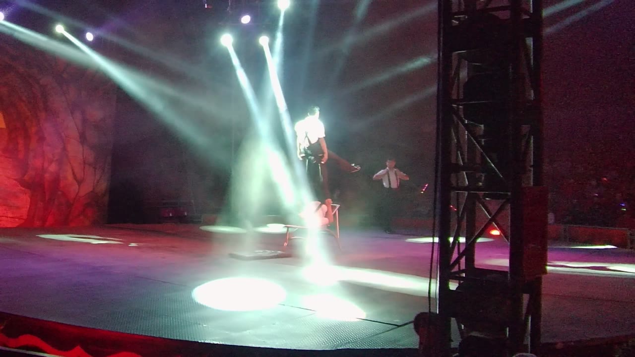 In San Jose Del Cabo, the acrobat rises into the white spotlights while a second performer stands to the right and the red scenic backdrop glows behind them.