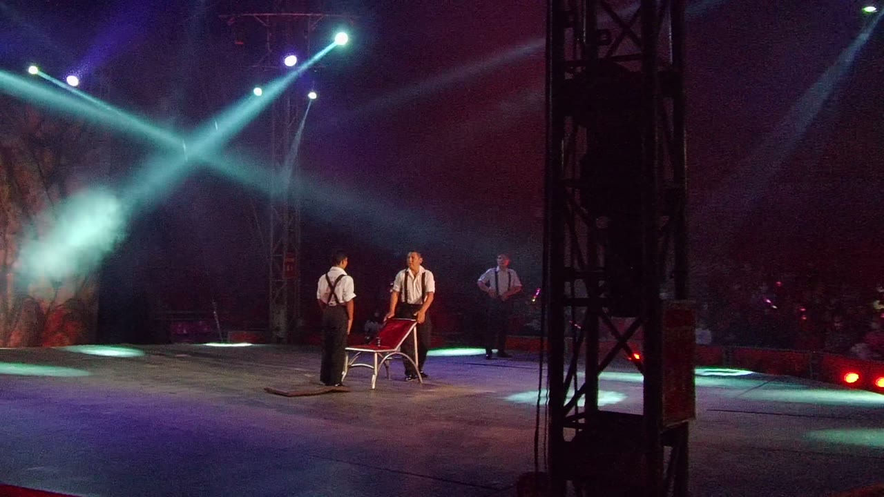 In San Jose del Cabo, Mexico, stage performers in white shirts and red suspenders stand around a red chair as crossed spotlights cut through smoke above the audience.