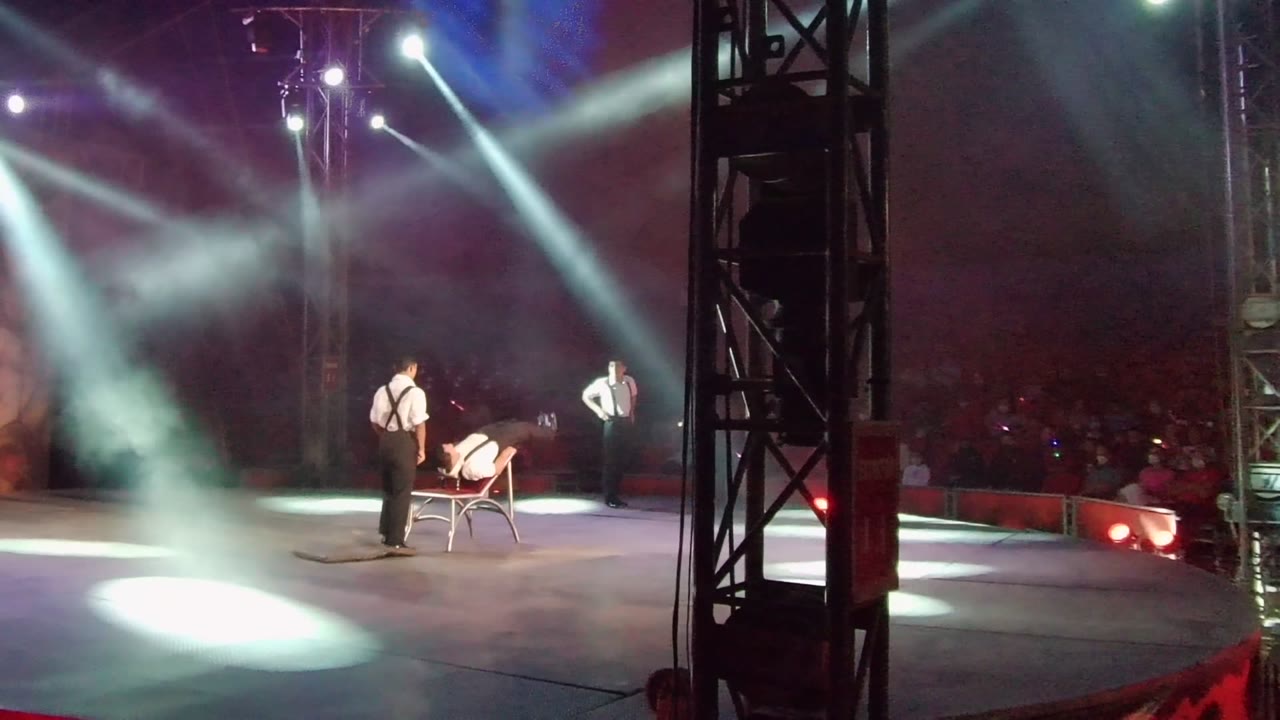 In San Jose del Cabo, Mexico, one performer leans over the red chair as two other performers stand nearby, with diagonal spotlights and smoke crossing the dark stage.