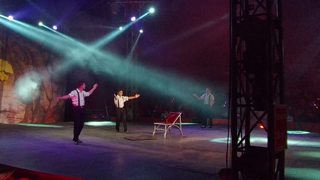 Three performers on a San Jose del Cabo stage gesture under blue-white spotlights, with a red chair prop and rock-like backdrop visible.