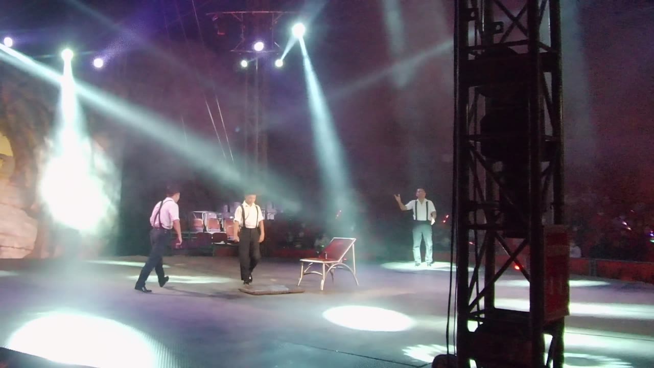 Two performers move near the red chair on the San Jose del Cabo stage while a third stands at right under intense white spotlight beams.