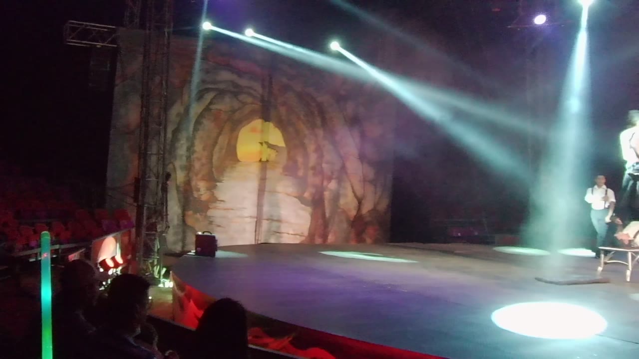 Wide view of a San Jose Del Cabo stage with bright spotlights, a cave-themed backdrop, a chair at right, audience silhouettes in front, and performers visible near the side of the set.