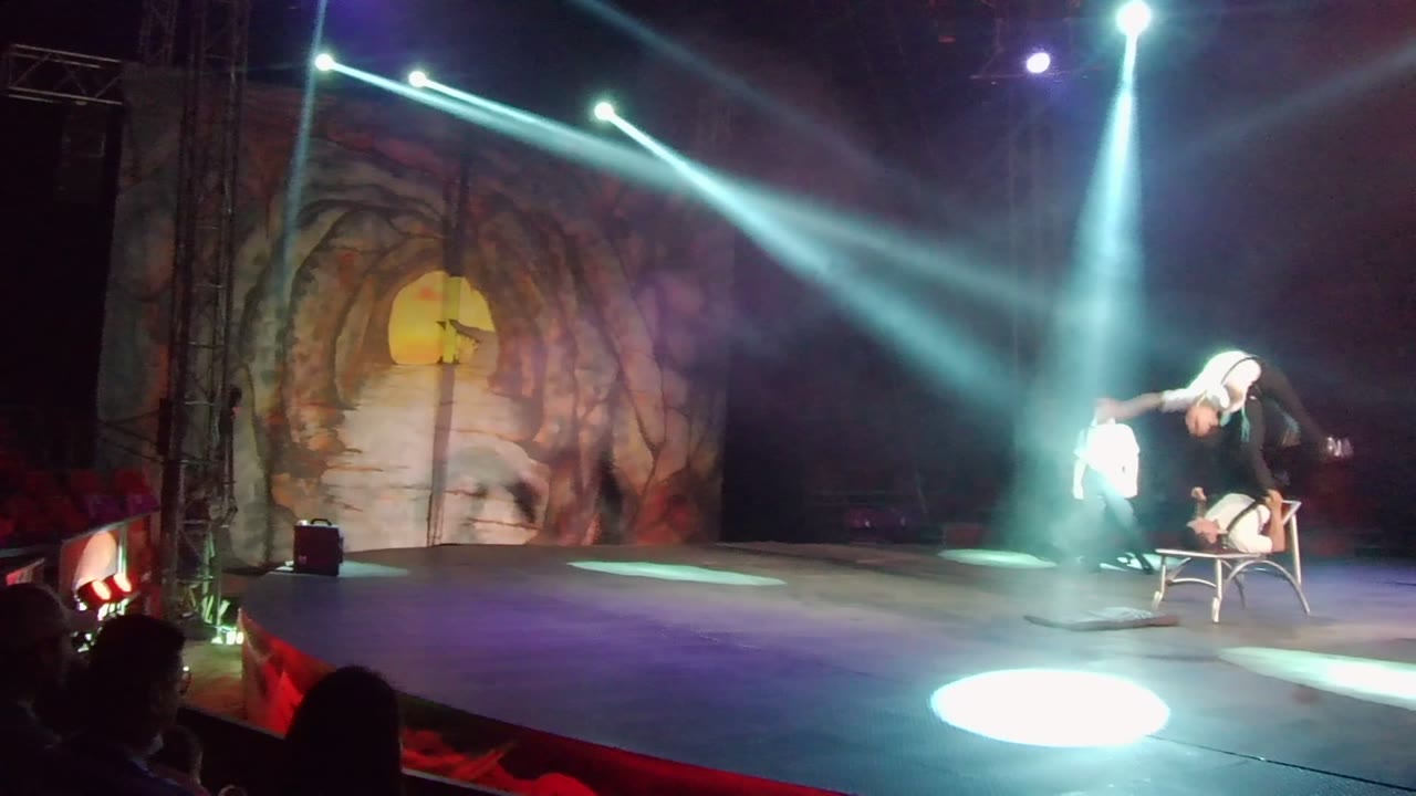Performer leans over a chair under crossing spotlights while another stands nearby