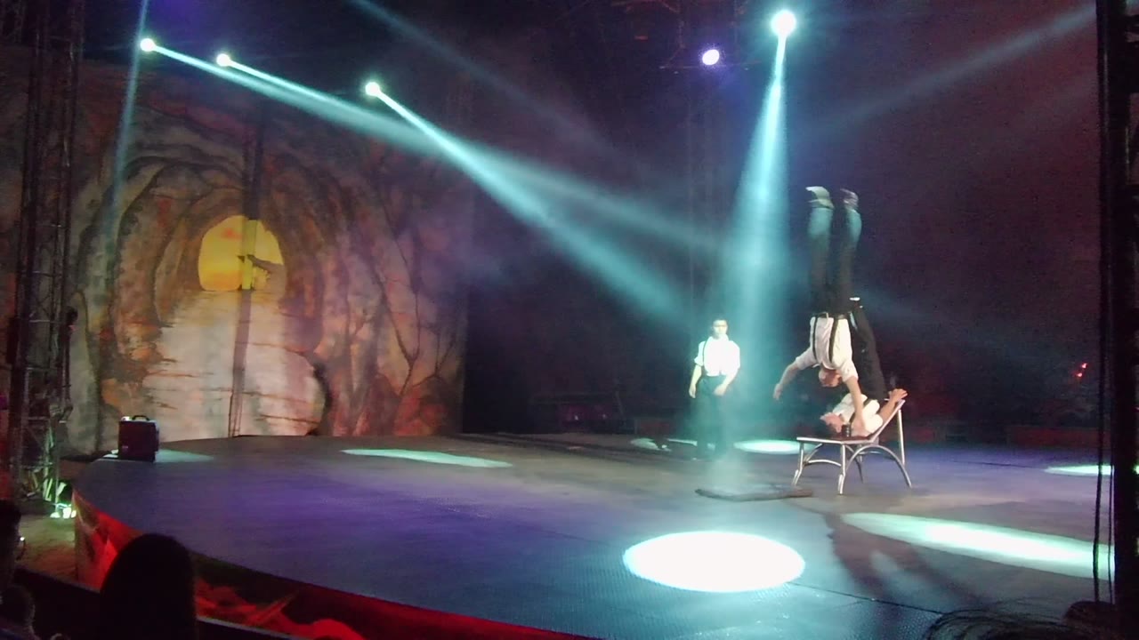 A performer holds an inverted handstand on a chair at right while another performer stands farther back on the San Jose Del Cabo stage beneath bright spotlights.