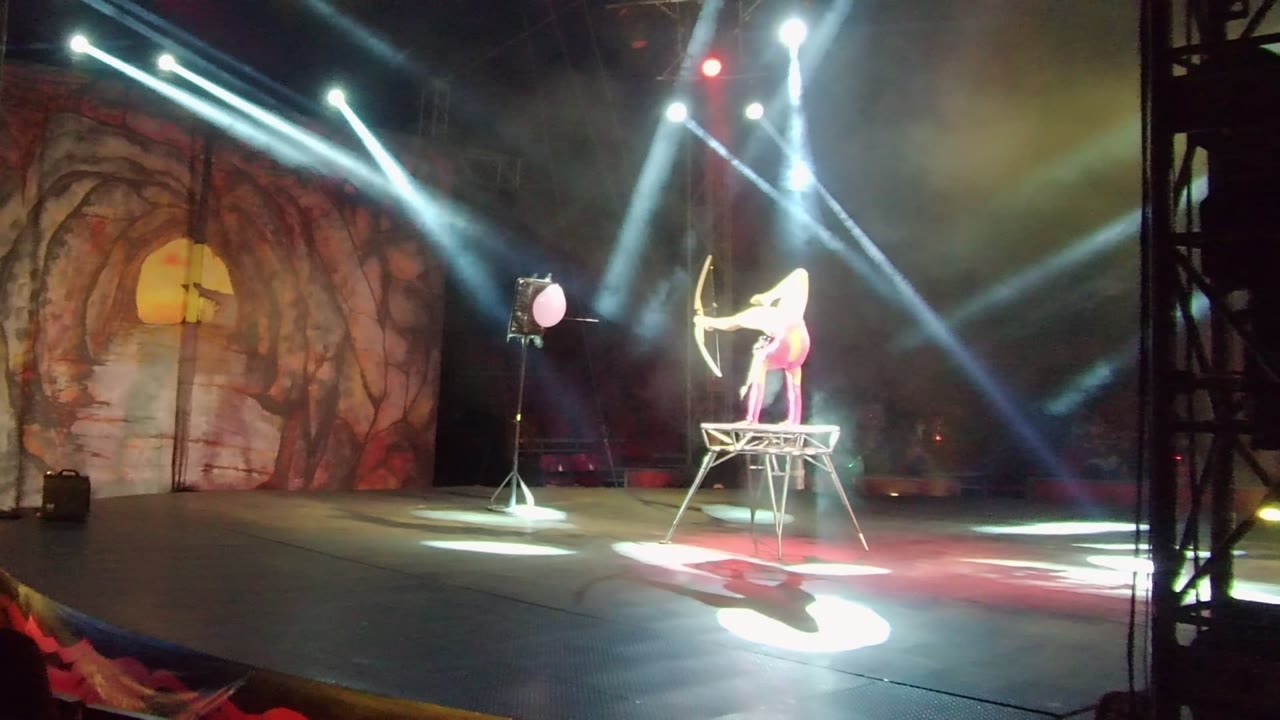 A wide view shows the performer on the platform at right of center, the bow catching bright light, and the cave-themed stage backdrop with warm red and yellow tones.