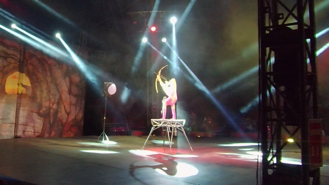 In San Jose del Cabo, Mexico, an acrobat stands on a raised metal platform holding a bow while white spotlights cross above a cave-themed stage backdrop.