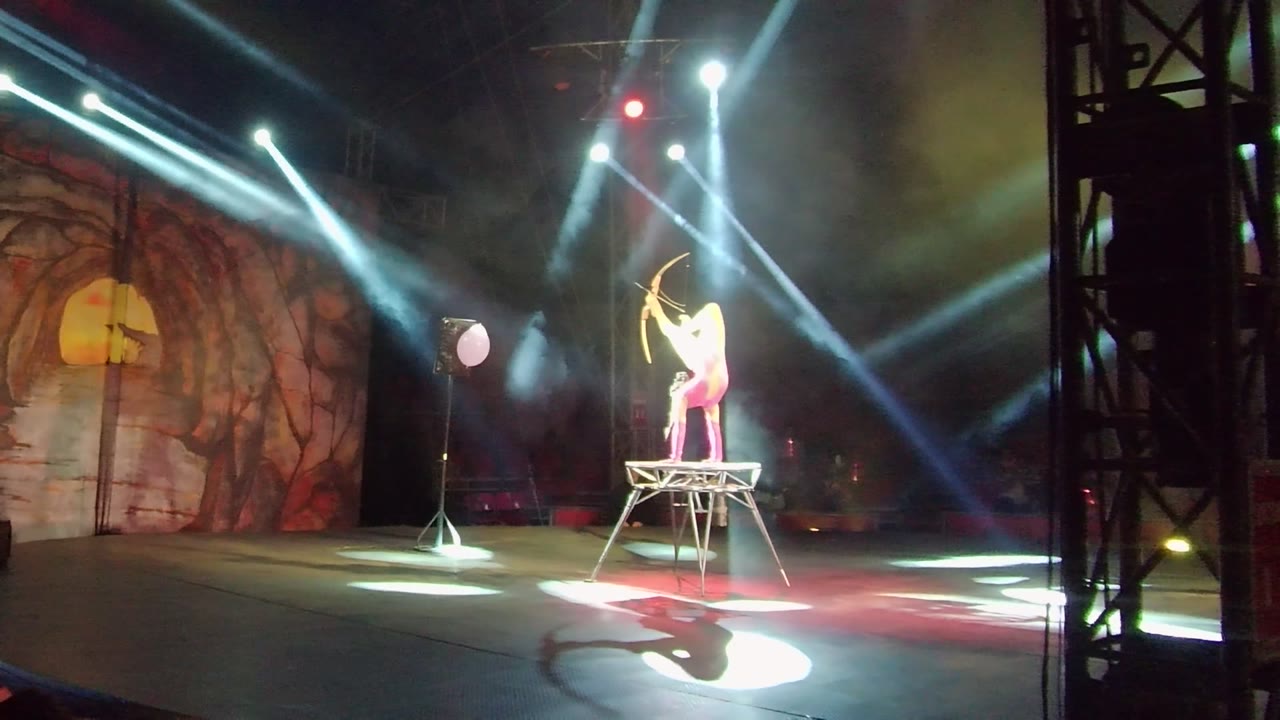 Performer aims a bow from a high platform on a dark stage.