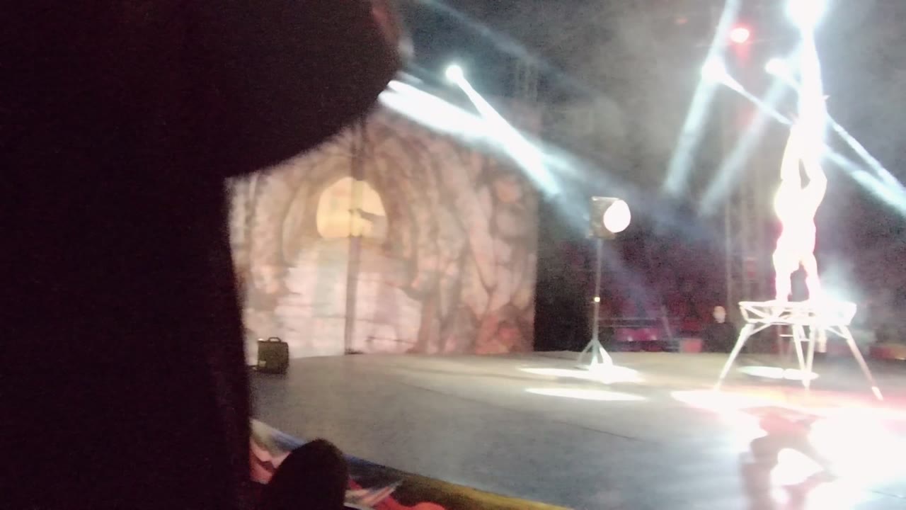 At 0:00 in San Jose Del Cabo, a dark foreground obstruction covers the left side while an acrobat stands on a platform under strong stage lights.