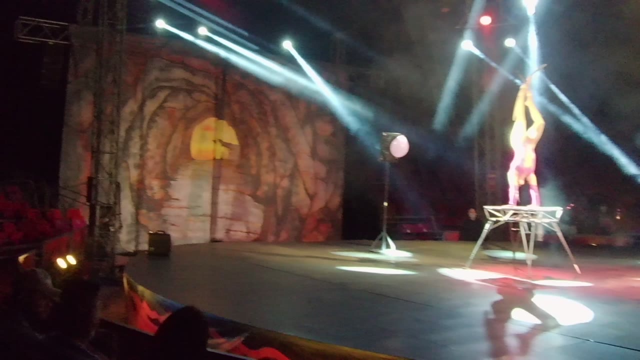 Acrobat stands on a platform with arms raised under spotlights and a painted backdrop.