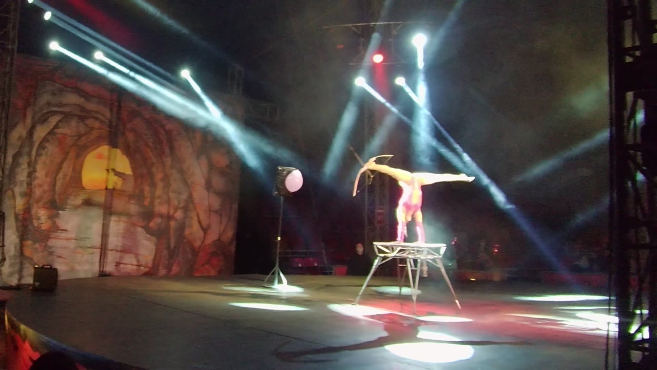 In San Jose del Cabo, Mexico, an acrobat extends into a split-like balance on a raised stool while bright beams cross a painted stage backdrop.