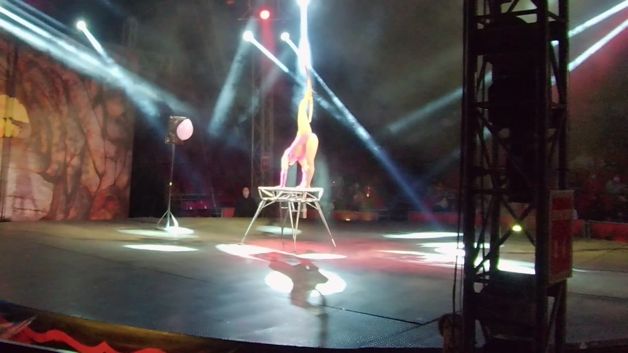 In San Jose del Cabo, a pink-costumed acrobat holds a vertical balance on a small table as white beams cut through stage smoke and a painted backdrop sits behind.
