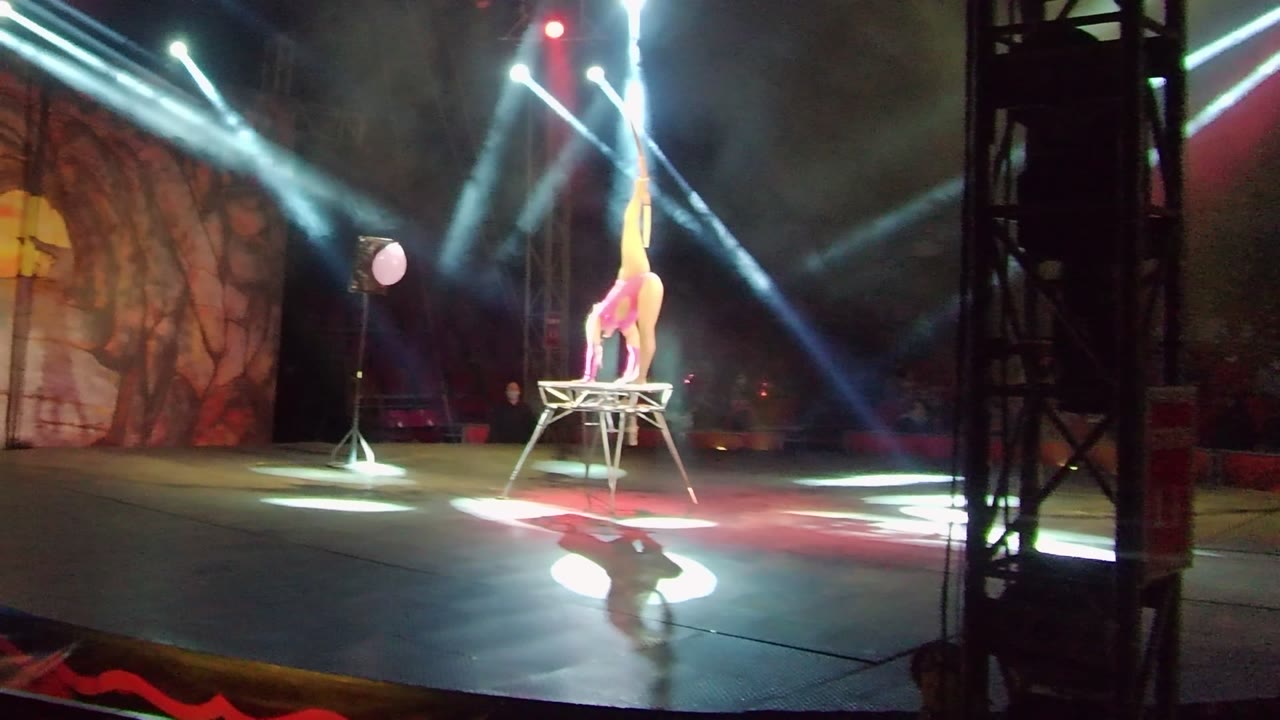 In San Jose del Cabo, the acrobat keeps a high vertical leg line on the table as bright beams rake across the smoky background and the open stage floor.
