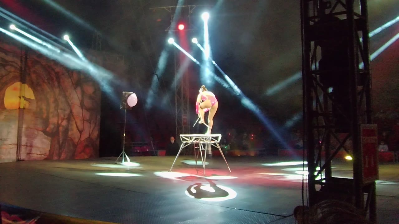 In San Jose del Cabo, an acrobat in a pink outfit crouches on a raised wire structure while white beams cross the stage and a mural backdrop fills the left side.