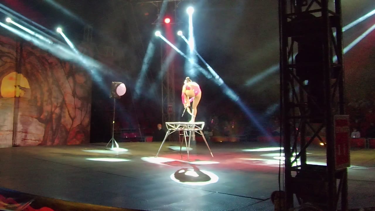 Acrobat balancing on raised wire structure with spotlights overhead.