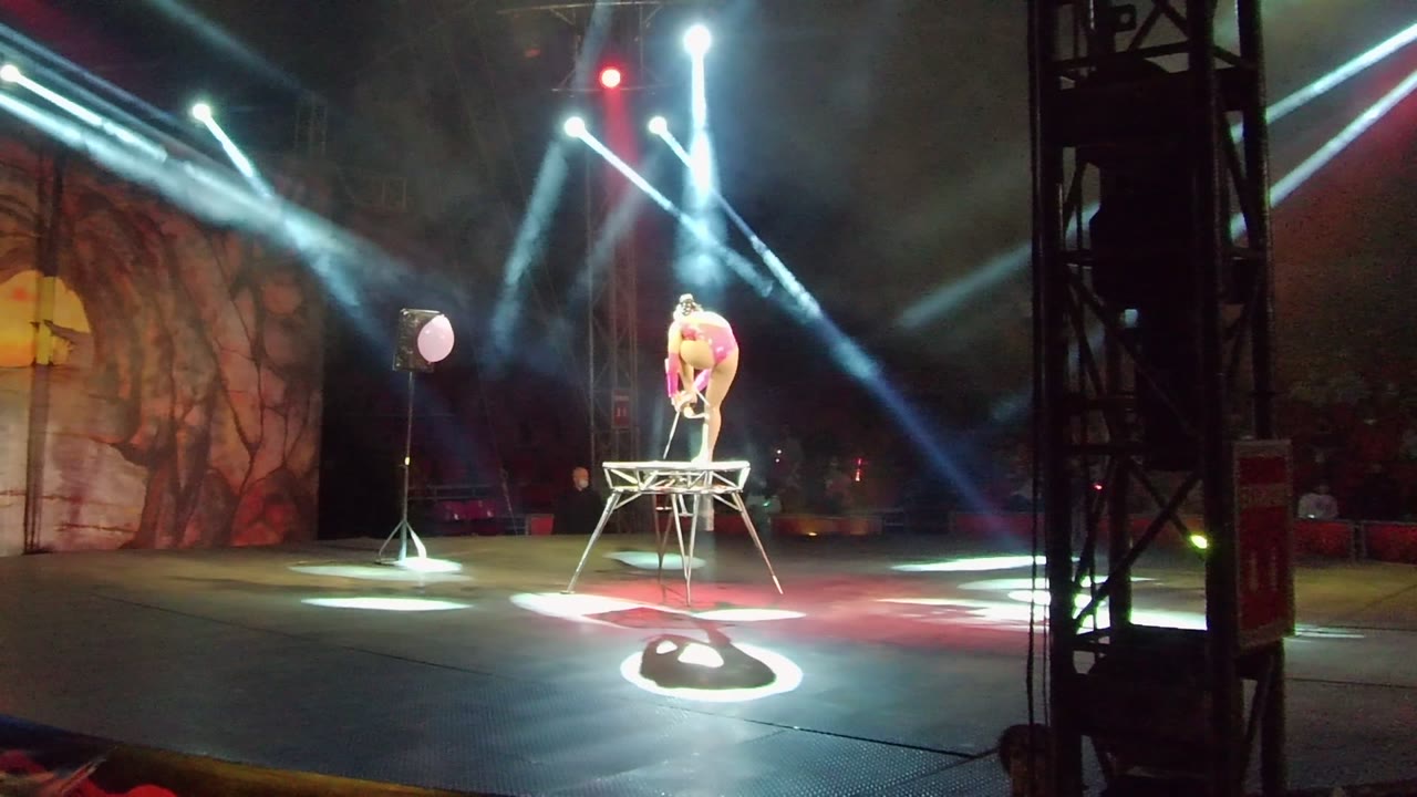 In San Jose del Cabo, the acrobat bends forward on the elevated wire structure as bright stage beams converge above and a circular spotlight marks the floor below.