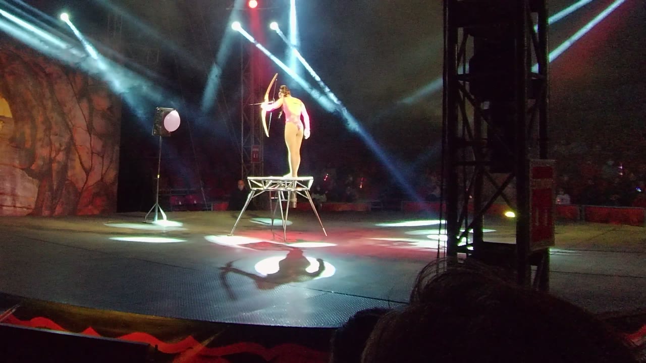 In San Jose Del Cabo, a circus performer stands on a raised metal platform and aims a bow toward a balloon target while blue-white spotlights cross the stage.