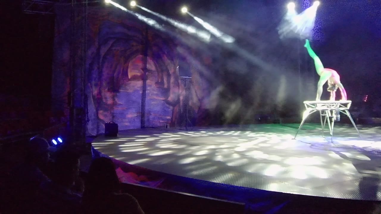 A performer in San Jose Del Cabo balances on a small stage stand at the right side of a smoky stage while audience silhouettes sit in the foreground.