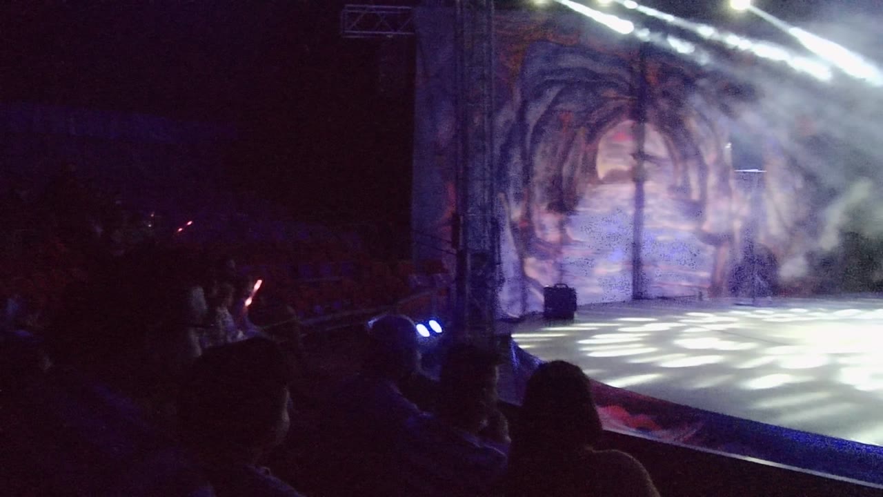 At 0:00 in San Jose del Cabo, Mexico, a dark theater stage is lit by white spotlights through smoke, with audience silhouettes in the foreground and a painted scenic backdrop behind the empty performance area.
