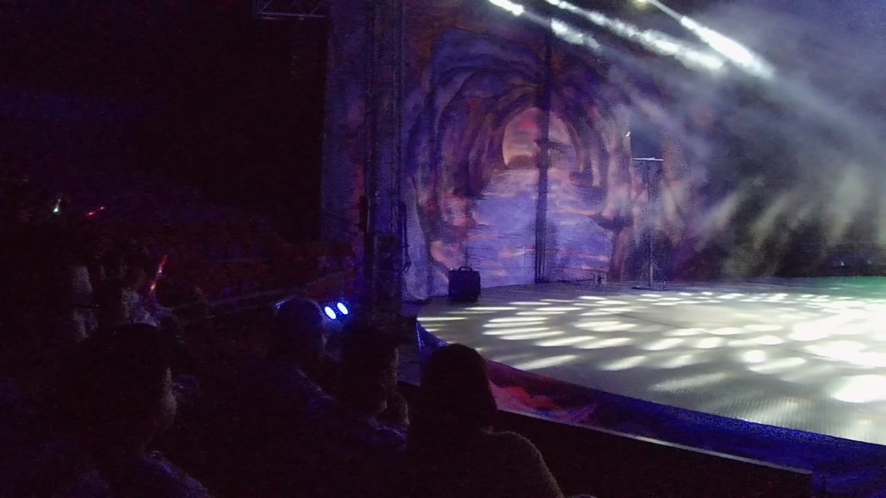 At 0:02 in San Jose del Cabo, Mexico, the stage floor catches patterned white light while smoke drifts below the overhead beams and the audience remains silhouetted at the front edge of frame.