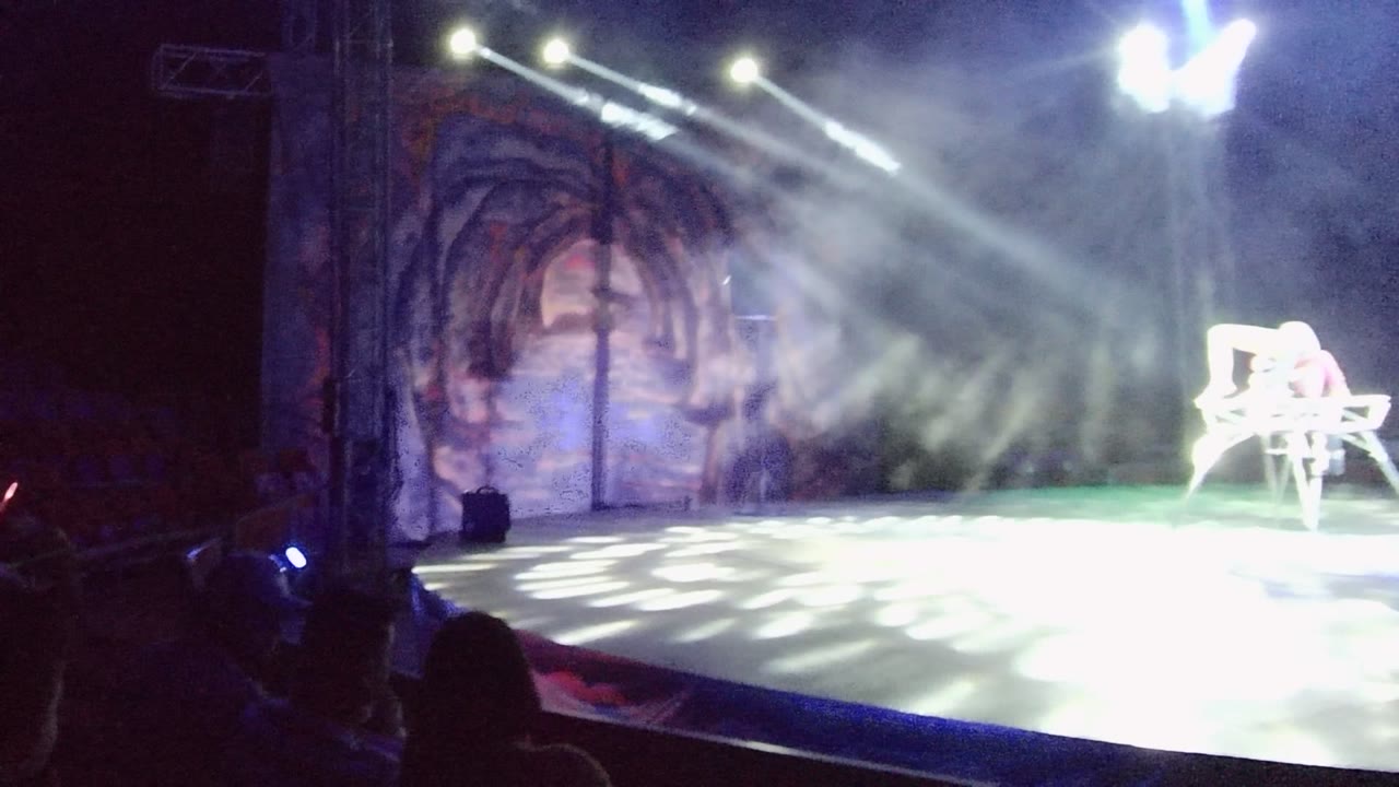 Wide audience-view frame of a San Jose Del Cabo stage with smoke, bright white beams, a cave-themed backdrop, a white chair at stage right, and silhouetted viewers in front.