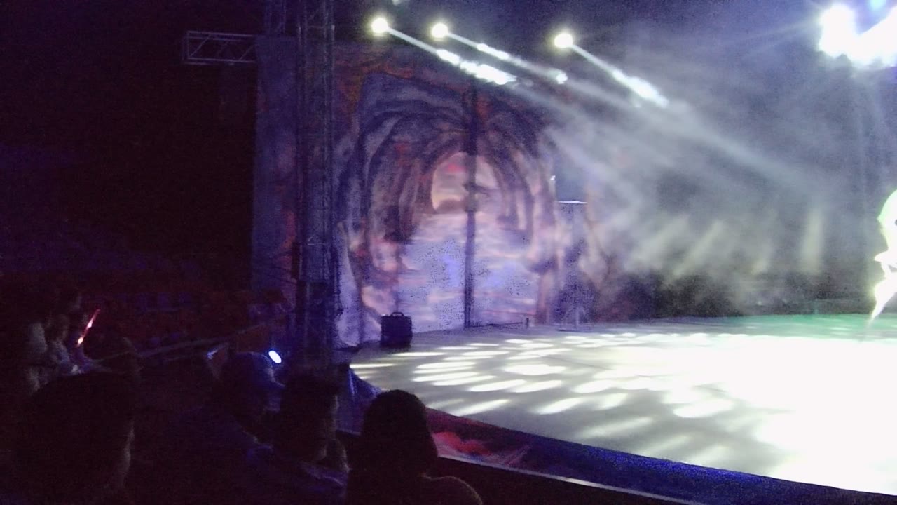 San Jose Del Cabo performance stage with the white chair partly exiting at the right edge, a cave-themed mural backdrop centered behind the haze, and audience shapes in shadow.