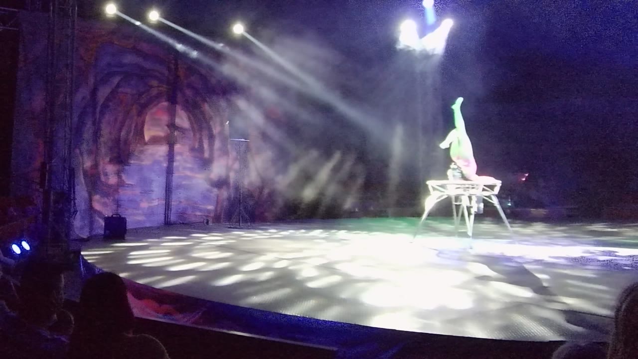 At 0:02 in San Jose del Cabo, Baja California Sur, Mexico, the acrobat keeps one leg extended high while balancing on the apparatus at the right side of a smoky, blue-lit stage.