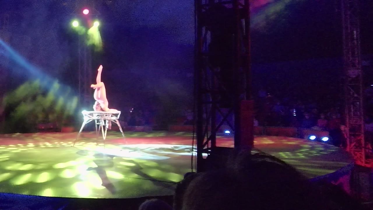 In San Jose Del Cabo, Baja California Sur, an acrobat balances upside down on a chair while green and purple light spreads across the stage and audience silhouettes sit in the foreground.