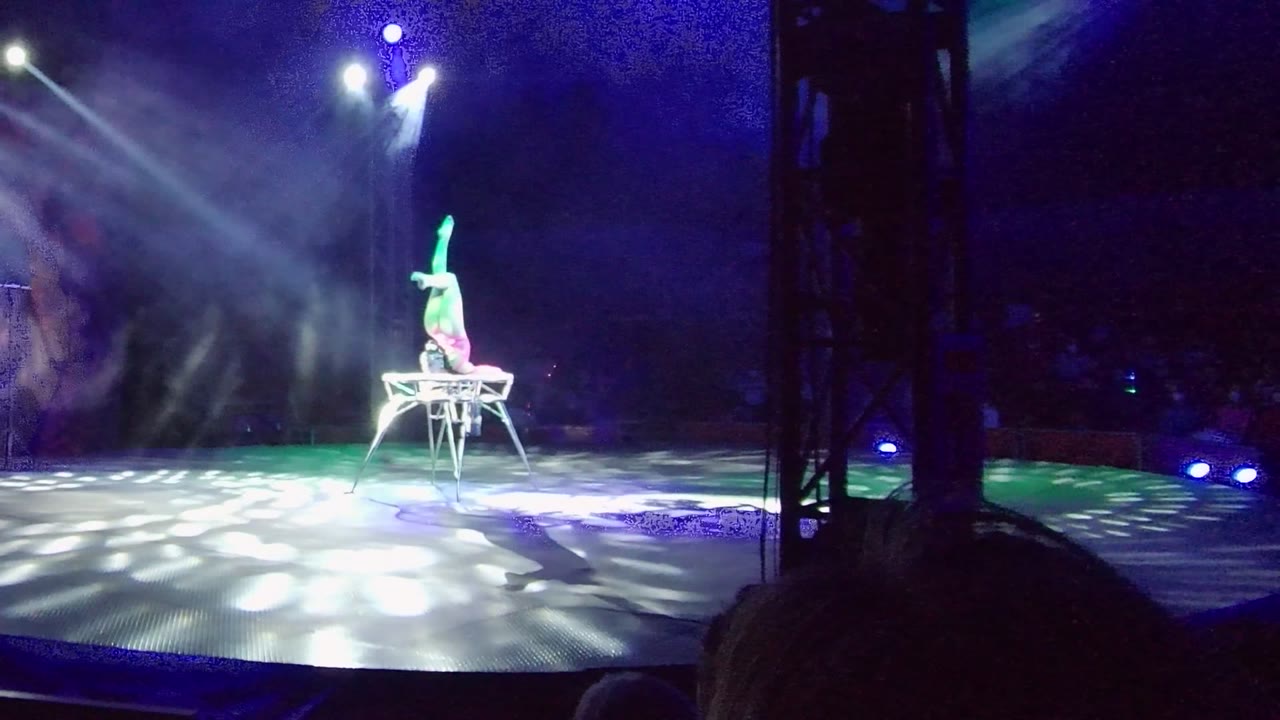 Acrobat continues an inverted pose on a chair in white stage light.