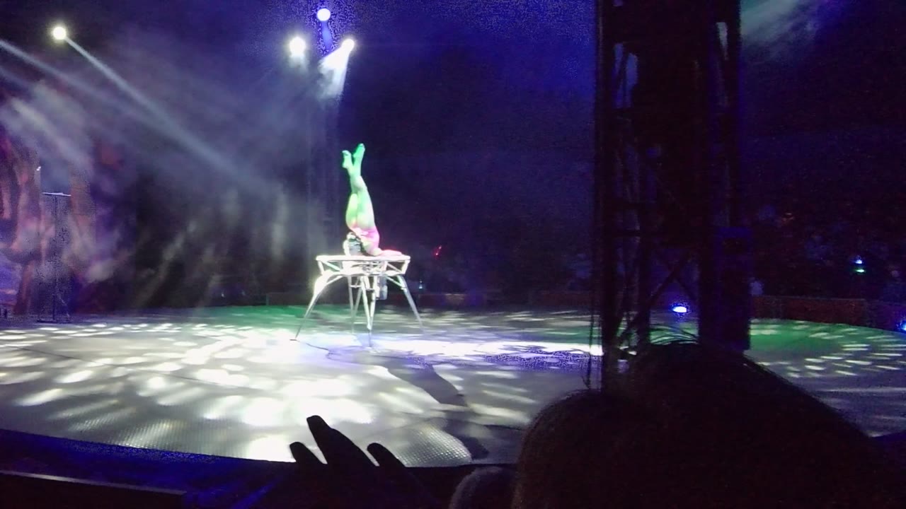 In San Jose Del Cabo, Baja California Sur, the acrobat's legs are lit green while held vertical above the chair, with audience hands and heads at the bottom edge and blue-white beams overhead.