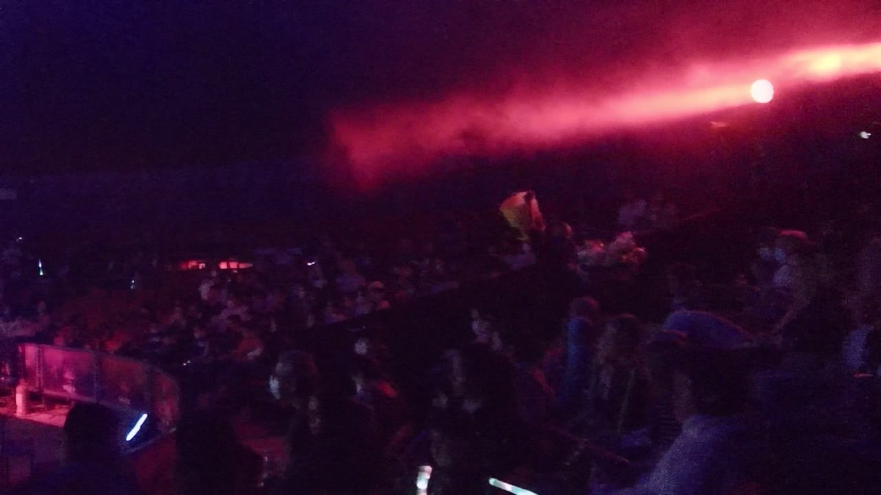 San Jose del Cabo nighttime concert crowd seen from near the stage edge, with red smoke overhead, purple lighting, and people visible in the dark audience area.