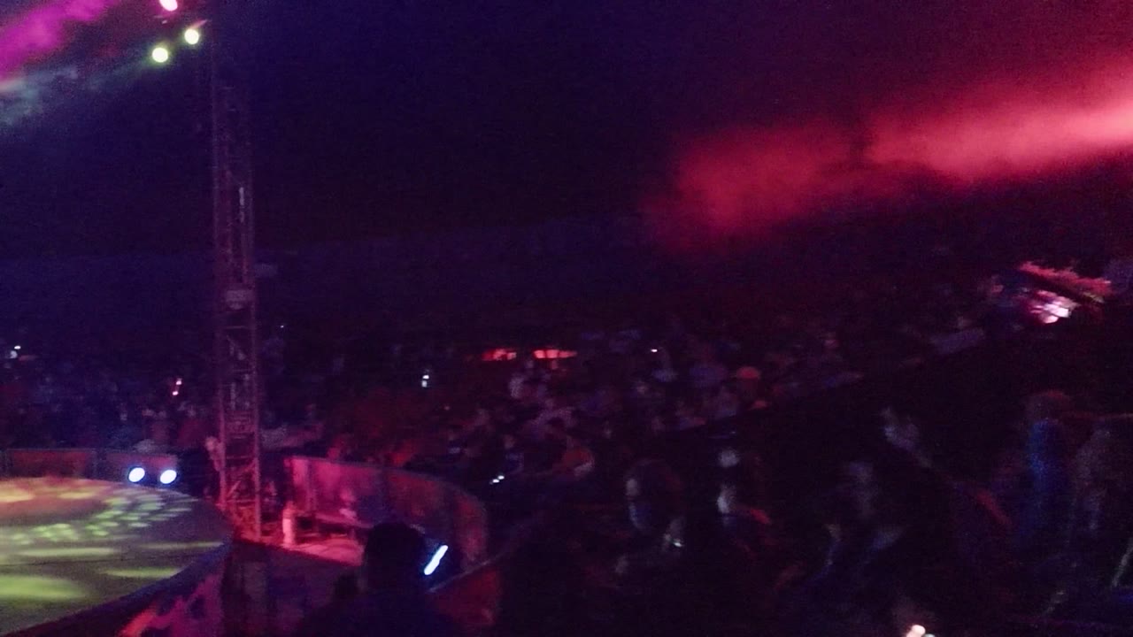San Jose del Cabo night concert view with the stage platform at left, audience in shadow, red smoke across the upper frame, and visible people near the stage edge.