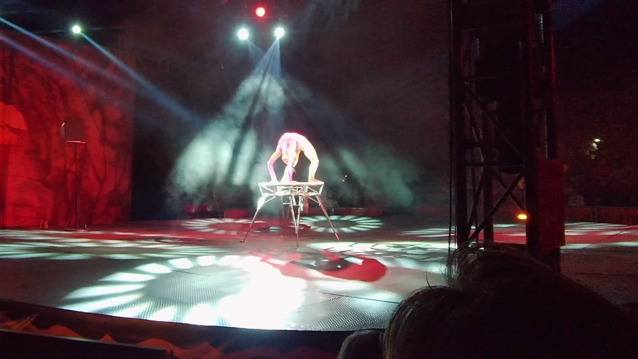 In San Jose Del Cabo, Mexico, a white-clad performer bends over a raised stage apparatus while red scenic panels, smoke, and bright spotlights frame the stage.