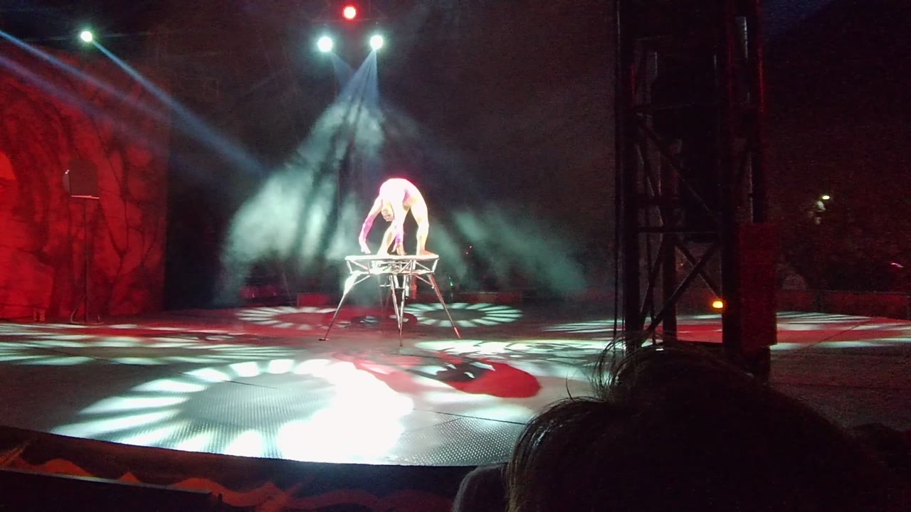 Performer remains bent over the apparatus with white spotlights cutting through smoke.
