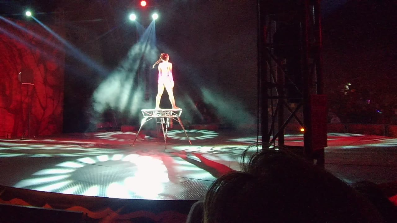 A white-clad performer stands upright on the raised apparatus beneath crossed white spotlights, with red stage scenery and audience silhouettes visible in San Jose Del Cabo.