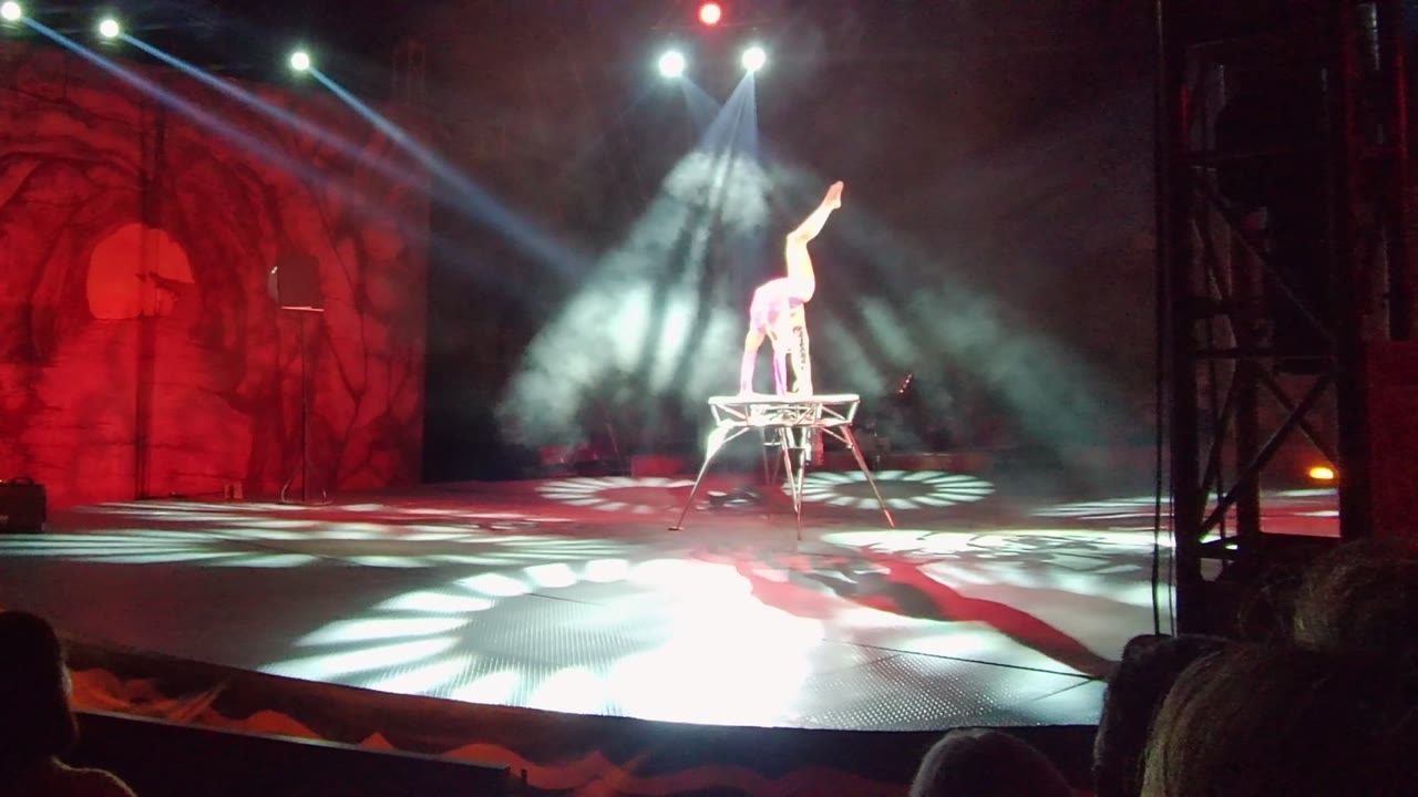 An acrobat in San Jose Del Cabo balances high on one hand and one leg on a small platform while white spotlights and red stage lighting fill the background.