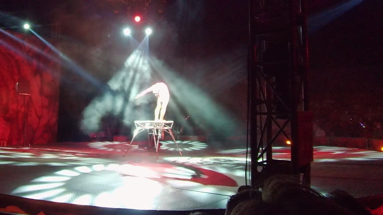 In San Jose Del Cabo, a white-clad performer bends forward on a raised metal platform while white spotlights and smoke fill a dark stage with a red backdrop.