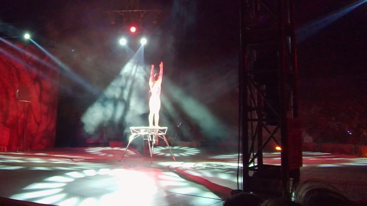 In San Jose Del Cabo, the performer remains centered on the platform with arms high while crossing white spotlights, haze, and red stage panels frame the pose.