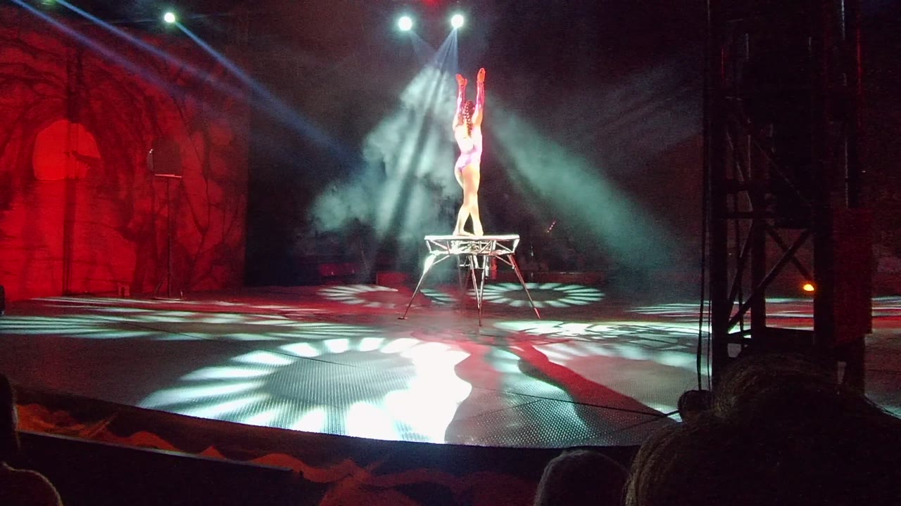 In San Jose del Cabo, the acrobat stands tall on the small platform with both arms raised as bright spotlights and floor patterns frame the stage.