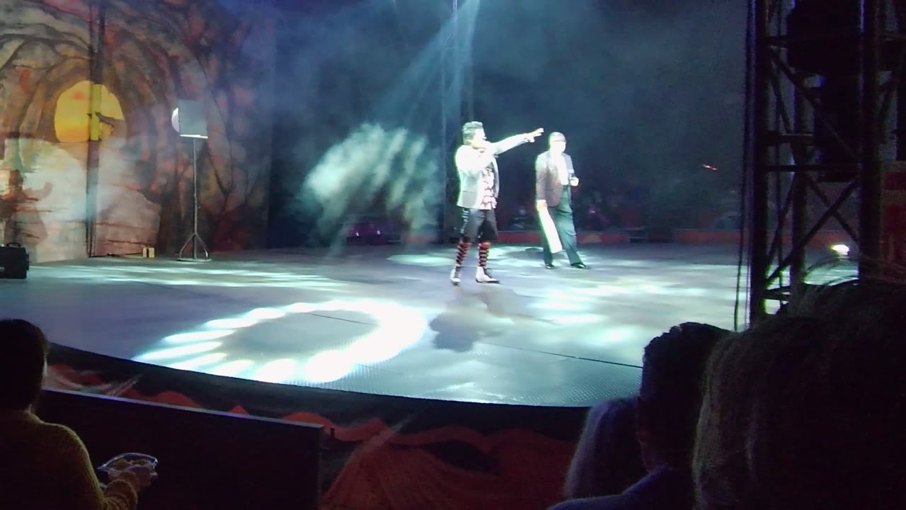 Performer gestures toward the side while another performer stands nearby on stage.